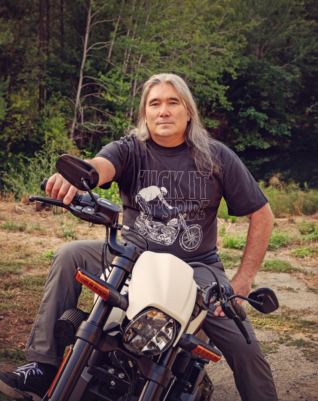 Man sitting on a motorcycle wearing an Iron & Velvet t-shirt with a graphic design, outdoors.