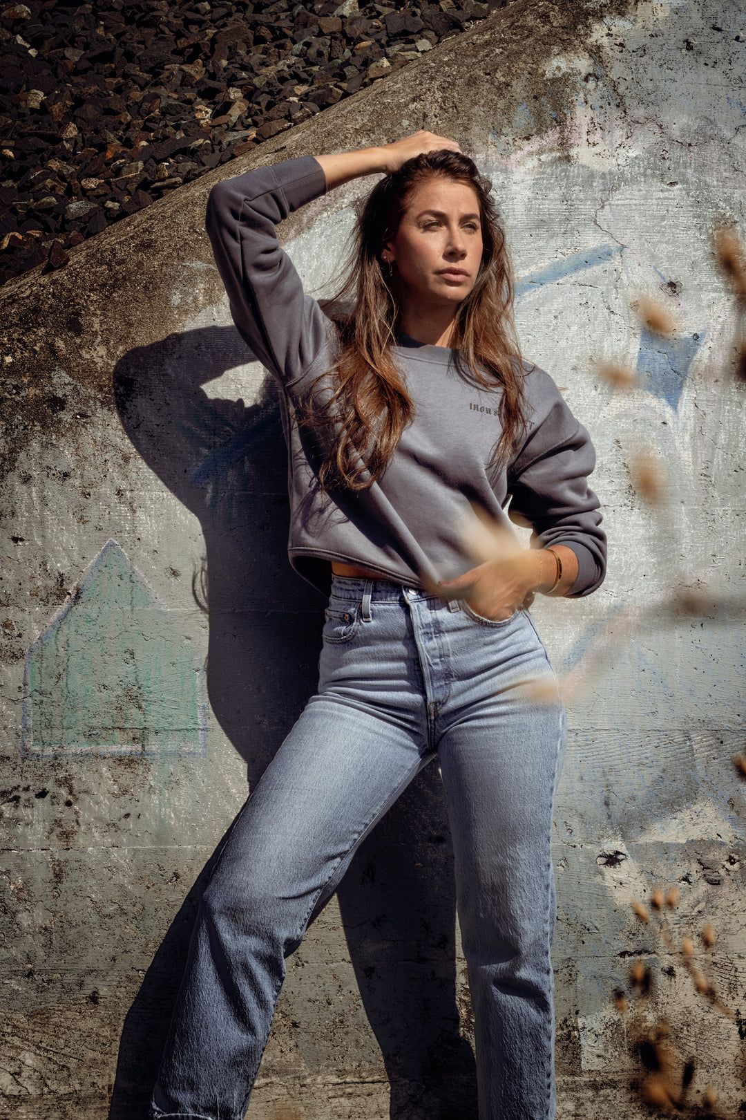 Woman in gray sweatshirt and blue jeans posing against a textured wall.